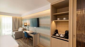 A hotel room with a bed, a desk with a chair, a TV mounted on the wall, a coffee maker, and a shelf with cups and a box.
