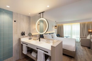 Quarto de hotel moderno com uma cama grande, banheiro separado com toucador, espelho redondo e vista para o exterior pela janela.