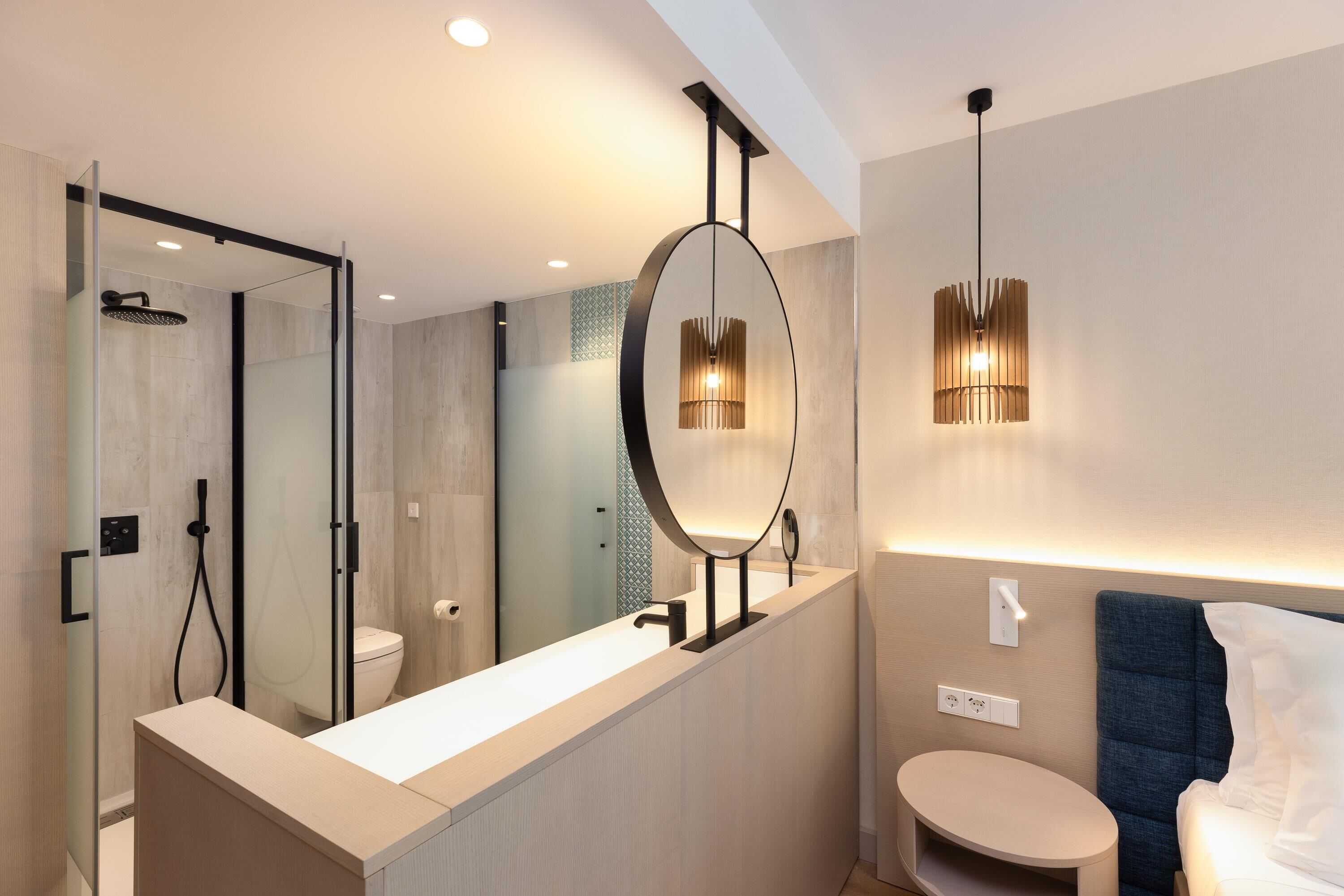Un baño moderno con ducha envuelta en vidrio, un espejo redondo, una mesita de noche y una cama con cabecera azul.