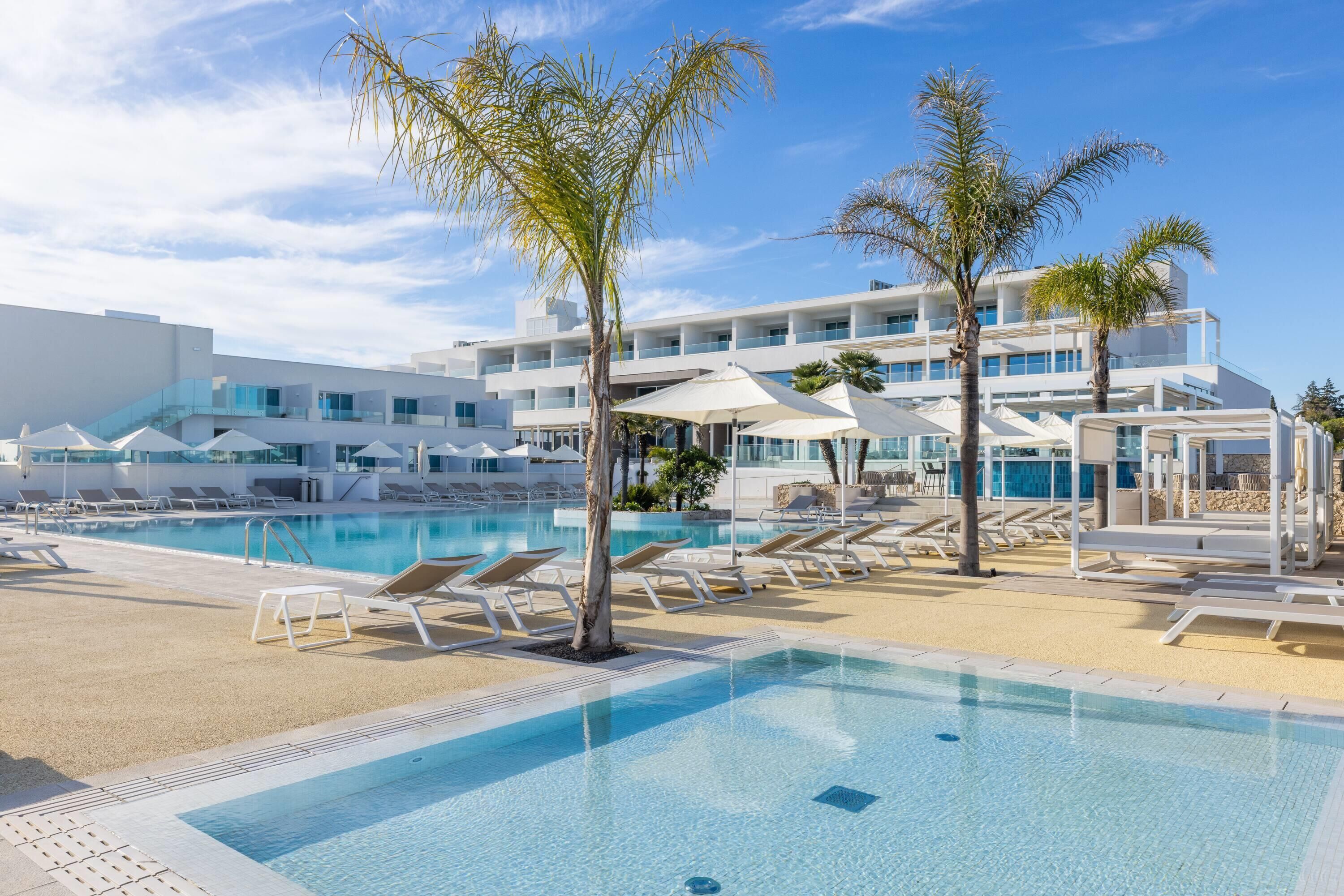 Une piscine avec des chaises longues et des palmiers, un bâtiment moderne doté de balcons, et un ciel dégagé.