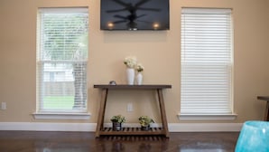 Una stanza con TV a parete, un comodino con oggetti decorativi e due finestre con tende.