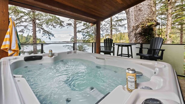 Een hottub met een blikje bier, omgeven door een houten terras, stoelen en uitzicht op bomen en water.
