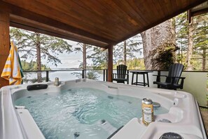 Un bain à remous, une bière à portée de main, entouré d’une terrasse en bois, de chaises et offrant une vue sur les arbres et l’eau.