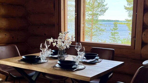 En matplats i en stuga med träpanel, ett bord dukat för två personer, en fällmatta av fuskpäls och utsikt över en sjö genom stora fönster.
