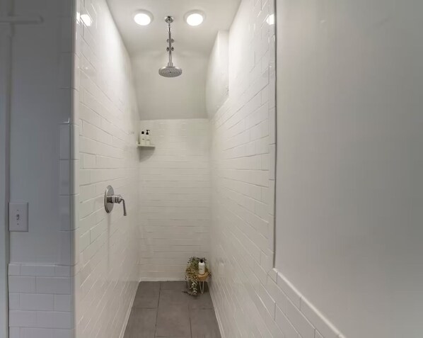 Un baño estrecho con paredes de azulejos blancos, un cabezal de ducha manual y un estante pequeño con artículos de aseo.