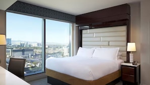 Une chambre d’hôtel avec un grand lit, un bureau, une chaise, une table de chevet et une vue sur le paysage urbain, visible à travers de grandes fenêtres.