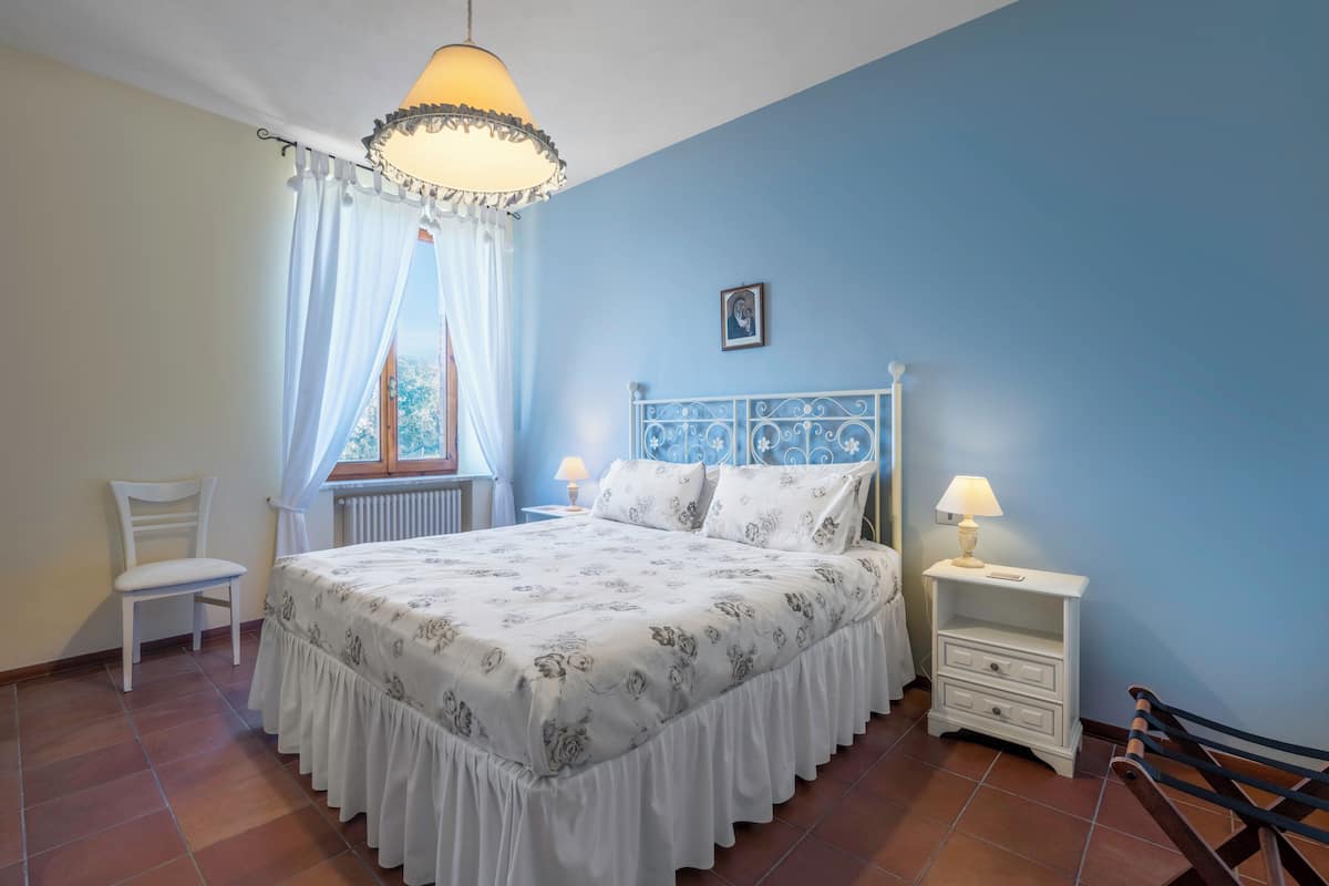 Un dormitorio con cama, una silla, una mesita de noche y un ventanal con cortinas.