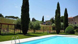 Una piscina con plataforma de madera, rodeada de altos cipreses y un edificio de piedra al fondo.