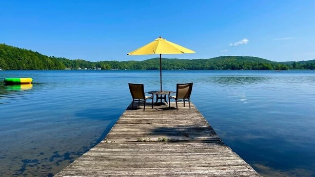 Une jetée en bois s’avance sur un lac paisible, avec un parasol jaune et deux chaises à son extrémité.