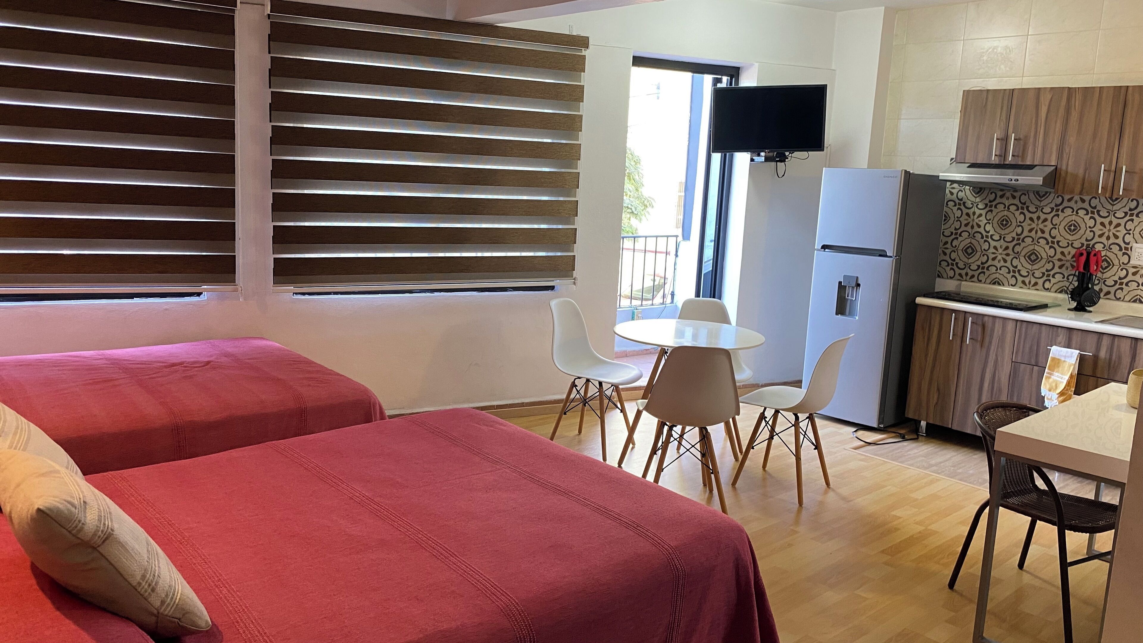 Une chambre avec deux lits, une petite table à manger avec des chaises, un coin cuisine équipé d’un réfrigérateur et un téléviseur fixé au mur.