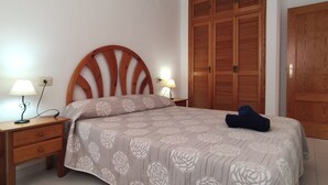 A bedroom with a wooden headboard, a bed with a floral bedspread, two bedside lamps, a wooden wardrobe, and a wooden door. - Apartamento Picasso 3 Inmomart (Peñiscola)