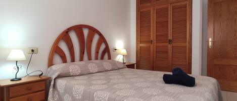 A bedroom with a wooden headboard, a bed with a floral bedspread, two bedside lamps, a wooden wardrobe, and a wooden door.
