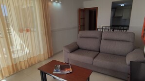 A living room with a sofa, a coffee table with magazines, and a kitchen area visible through an open door.