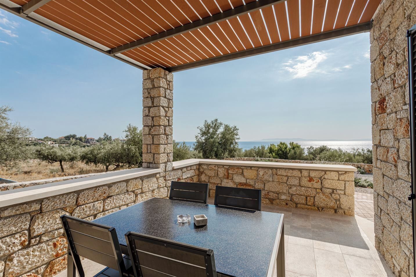 A stone patio with a table and chairs, overlooking a scenic view of the sea and hills.