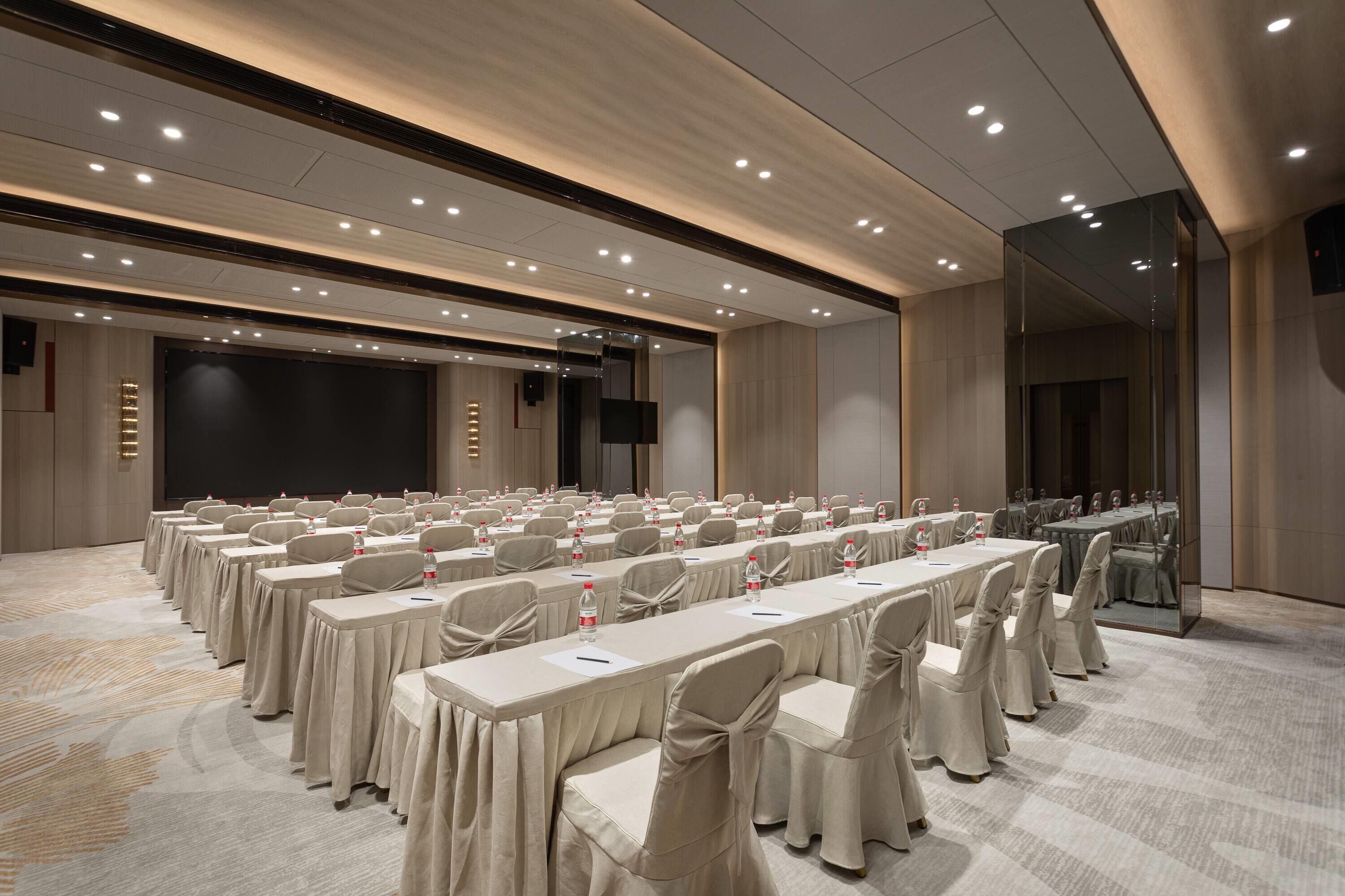 A conference room with rows of tables and chairs, a large screen, and a stage.