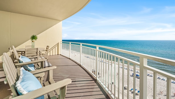 A balcony with wooden chairs and a table, overlooking a beach with a white railing and a clear view of the ocean. - Open Oct 25–Nov 2! Beautiful Condo Near Pier Park! (Panama City Beach)