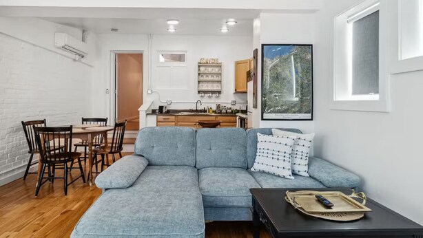 A modern living room with a sectional sofa, a dining table with chairs, a kitchen area, and a large window with a view of trees. - Cozy 1BDR APT in Vibrant Downtown Geneva (Geneva)
