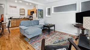 A modern living room with a sectional sofa, a dining area with a table and chairs, a kitchen with wooden cabinets, and a large window. - Cozy 1BDR APT in Vibrant Downtown Geneva (Geneva)