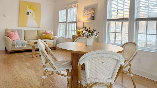 A spacious living room with a round wooden table, white wicker chairs, a beige sofa with pink and yellow cushions, and large windows with blinds.