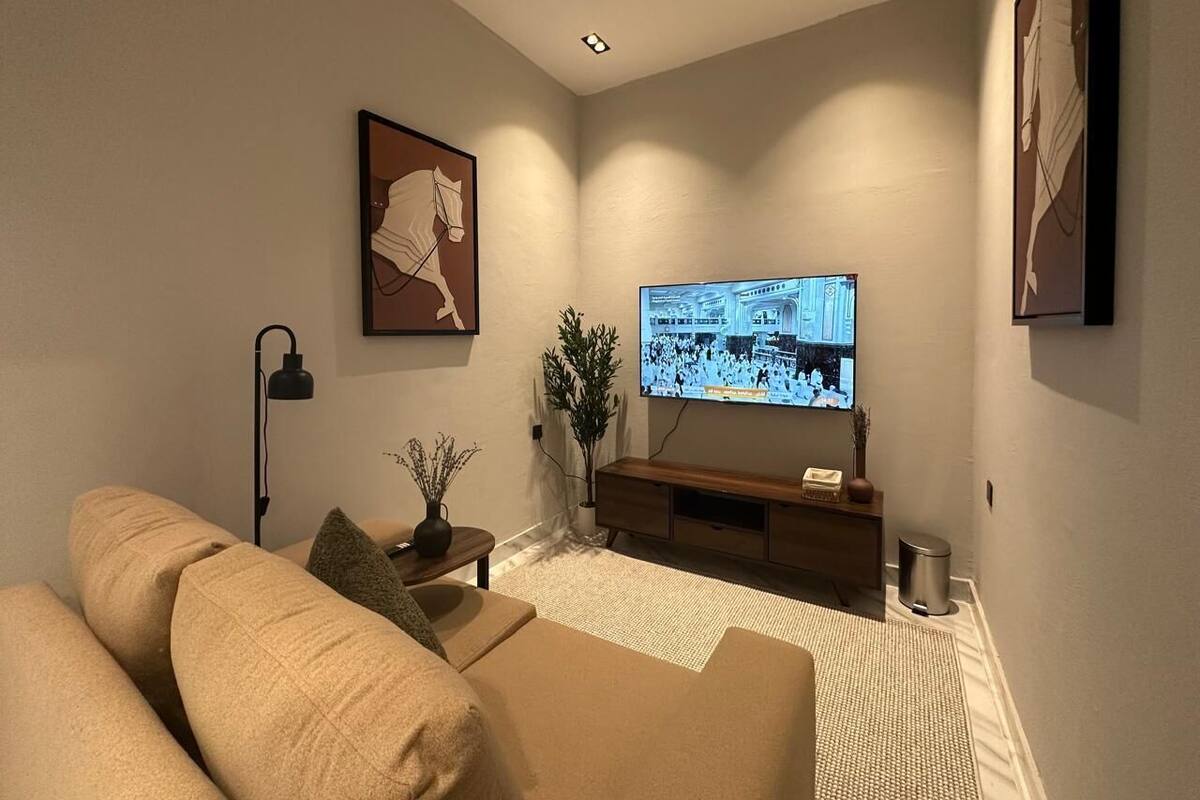 A modern living room with a beige sofa, a flat-screen TV mounted on the wall, a small side table with a lamp, and a decorative plant.