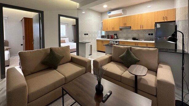 A modern living room with two beige sofas, a coffee table, and a small side table. A kitchen with wooden cabinets and a refrigerator is visible in the background. - M-Residence by Dayf Self-Service Apart. (Jeddah)