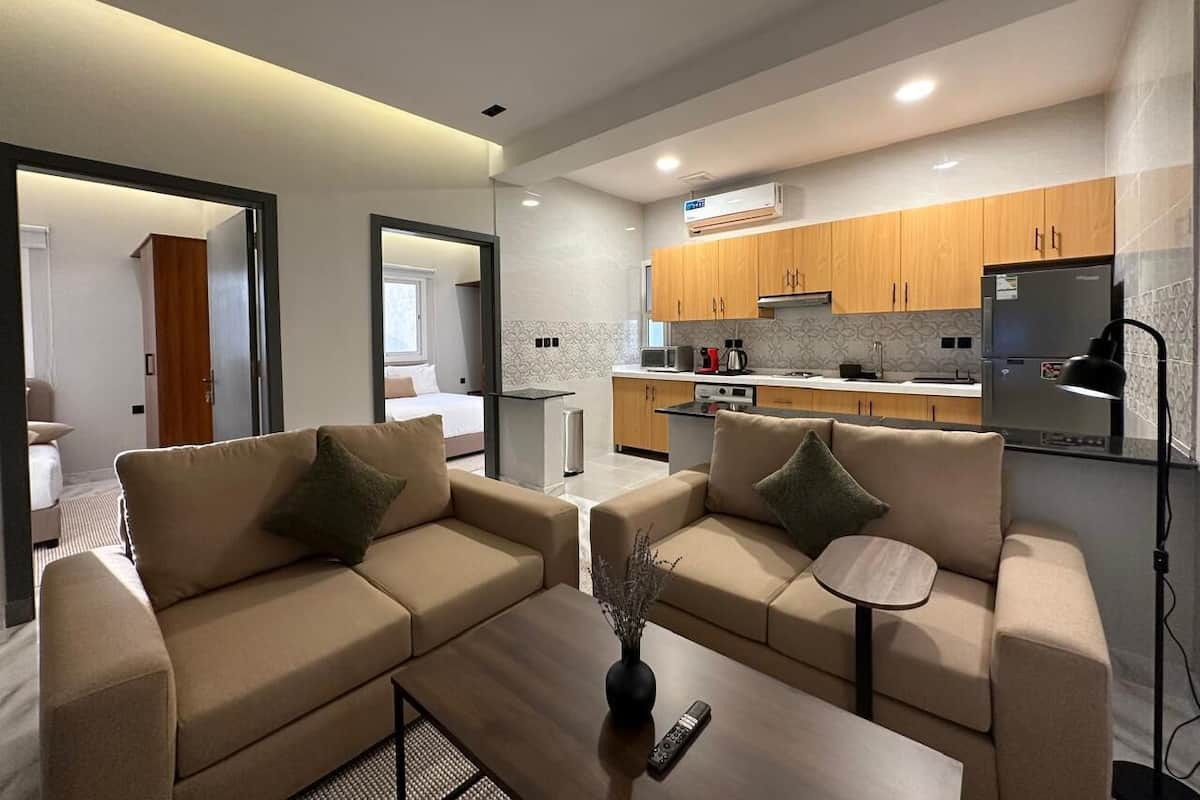 A modern living room with two beige sofas, a coffee table, and a small side table. A kitchen with wooden cabinets and a refrigerator is visible in the background.
