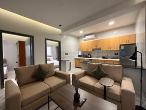 Un soggiorno moderno con due divani color beige, un tavolino da caffè e un piccolo tavolino laterale. Si intravede in secondo piano una cucina con mobili in legno e un frigorifero.