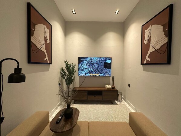 A modern living room with a flat-screen TV mounted on the wall, a wooden entertainment unit, a small plant, and two framed artworks on the walls. - M-Residence by Dayf Self-Service Apart. (Jeddah)