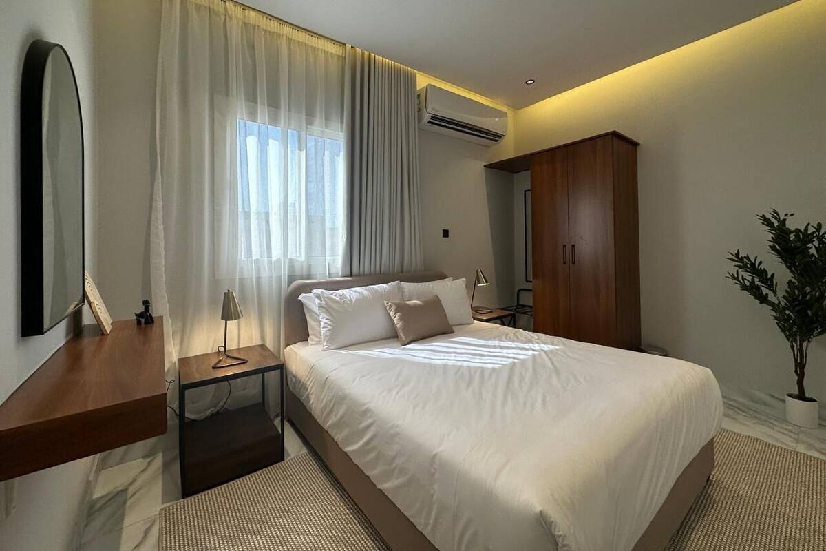 A modern hotel room with a large bed, a bedside table with a lamp, a wardrobe, and a potted plant.
