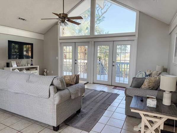 A spacious living room with a high ceiling, a ceiling fan, a large window, a television, a white sofa, a coffee table, and a fireplace.