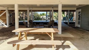 Um alpendre externo coberto com uma mesa e cadeiras de madeira, cercado por pilares de concreto.