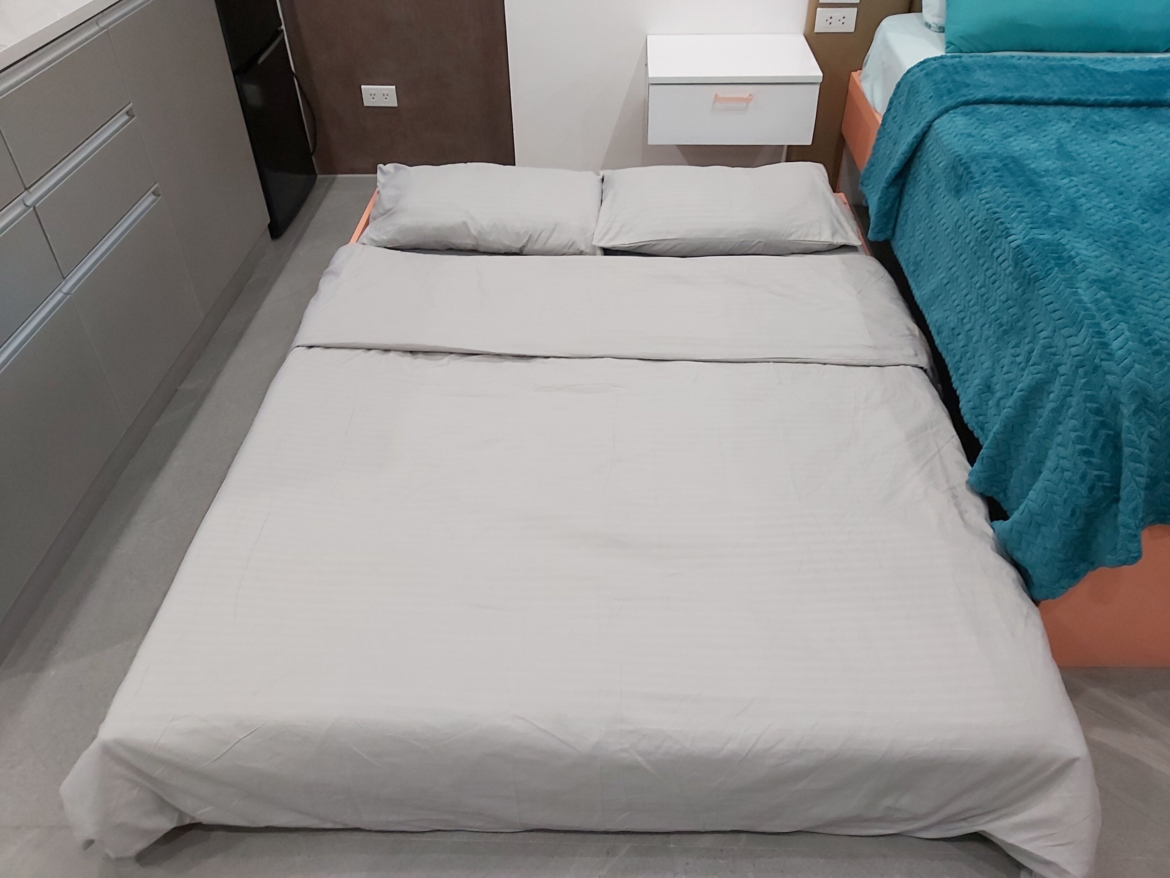 A single bed with a white mattress and pillows, a teal blanket, and a white bedside table with a drawer.
