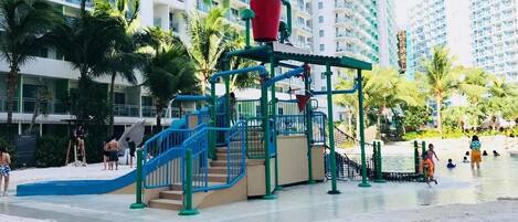 A resort area with a water play structure, lounge chairs, and high-rise buildings in the background.
