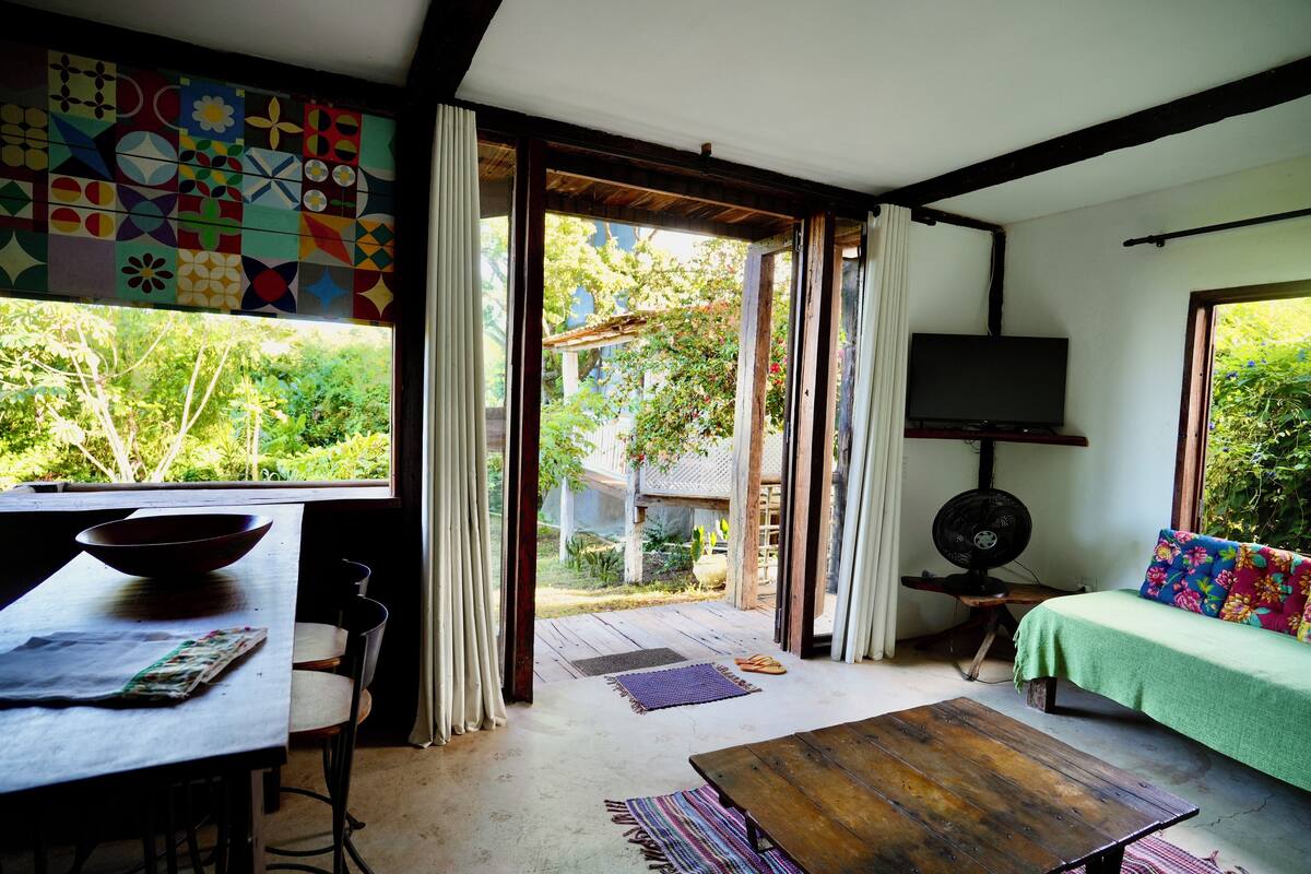 Ein Zimmer mit Holztisch, einer Couch mit farbigem Kissen, einem an der Wand montierten Fernseher und Blick ins Grüne.