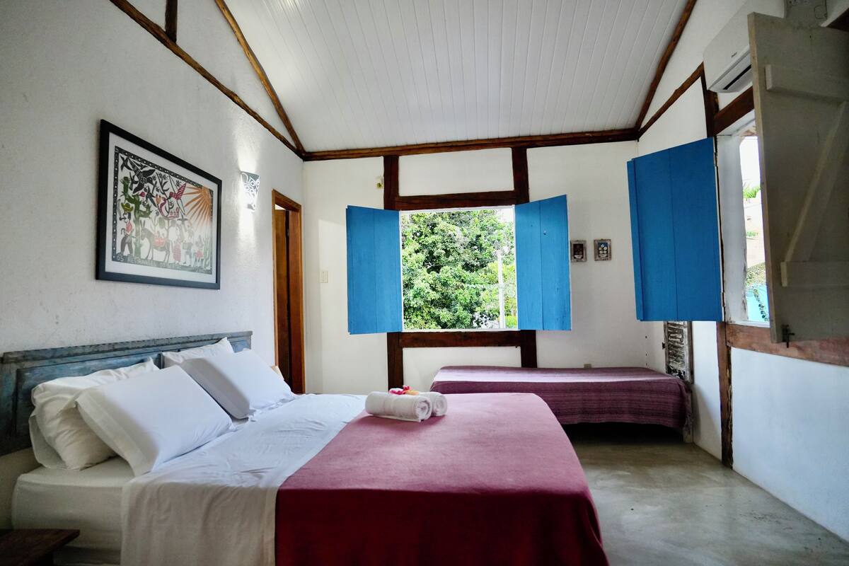 Ein Zimmer mit zwei Betten, eines mit weißem Kopfteil und das andere mit rotem, einem gerahmten Bild an der Wand und einem Fenster mit blauen Fensterläden.