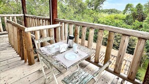 Een houten terras met een gedekte tafel voor twee personen, een koffiepot en uitzicht op een weelderig groen landschap.