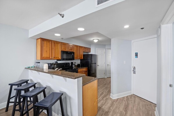 A modern kitchen with wooden cabinets, a black refrigerator, and a granite countertop. There are three black bar stools and a white door in the background. - Perdido Sun 800 (Pensacola)