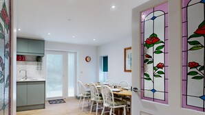 A dining area with a wooden table and white chairs, a kitchen with grey cabinets, and a door with stained glass featuring red roses.
