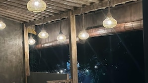 Un comedor interior con mesas y sillas de madera, lámparas colgantes de mimbre y grandes ventanales que ofrecen vistas del cielo nocturno.