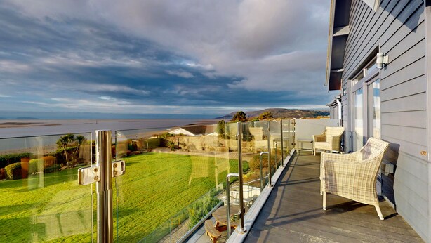 A balcony with wicker furniture overlooking a grassy yard and a body of water. - Waters Edge - Three Bedroom Cottage, Sleeps 6 (Deganwy)