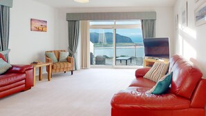 A living room with a red leather sofa, a striped armchair, a wooden coffee table, and a flat-screen TV. A large window offers a view of a coastal landscape. - Waters Edge - Three Bedroom Cottage, Sleeps 6 (Deganwy)