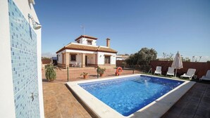 Une piscine avec une terrasse carrelée, un patio délimité par une clôture et, au fond, un bâtiment dont le toit est recouvert de tuiles.