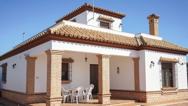 Une maison à deux étages, avec un toit en tuiles, des colonnes en briques et un patio équipé de chaises en plastique blanches.