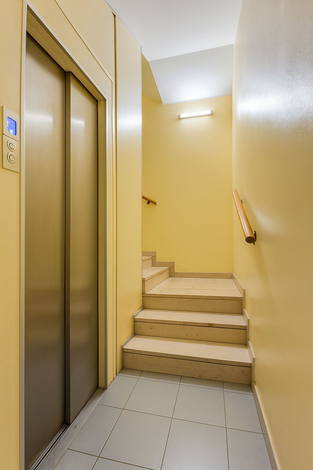 A narrow hallway with a beige elevator and a set of beige stairs leading to a landing.