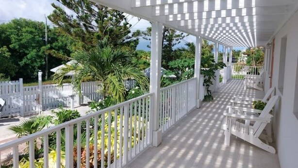 A covered outdoor area with white railings and chairs, surrounded by greenery and a view of the sea. - Buttonwood Lodge Unit #3 (Kew Town Settlement)