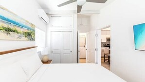 A bedroom with a large bed, white walls, a ceiling fan, and a painting of a beach scene. - Buttonwood Lodge Unit #3 (Kew Town Settlement)