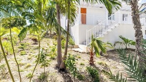 A white building with a staircase and a small porch area surrounded by palm trees and other vegetation. - Buttonwood Lodge Unit #3 (Kew Town Settlement)