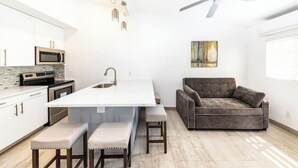 A modern kitchen with white cabinets, a central island with stools, a gray sofa, and a ceiling fan. - Buttonwood Lodge Unit #3 (Kew Town Settlement)