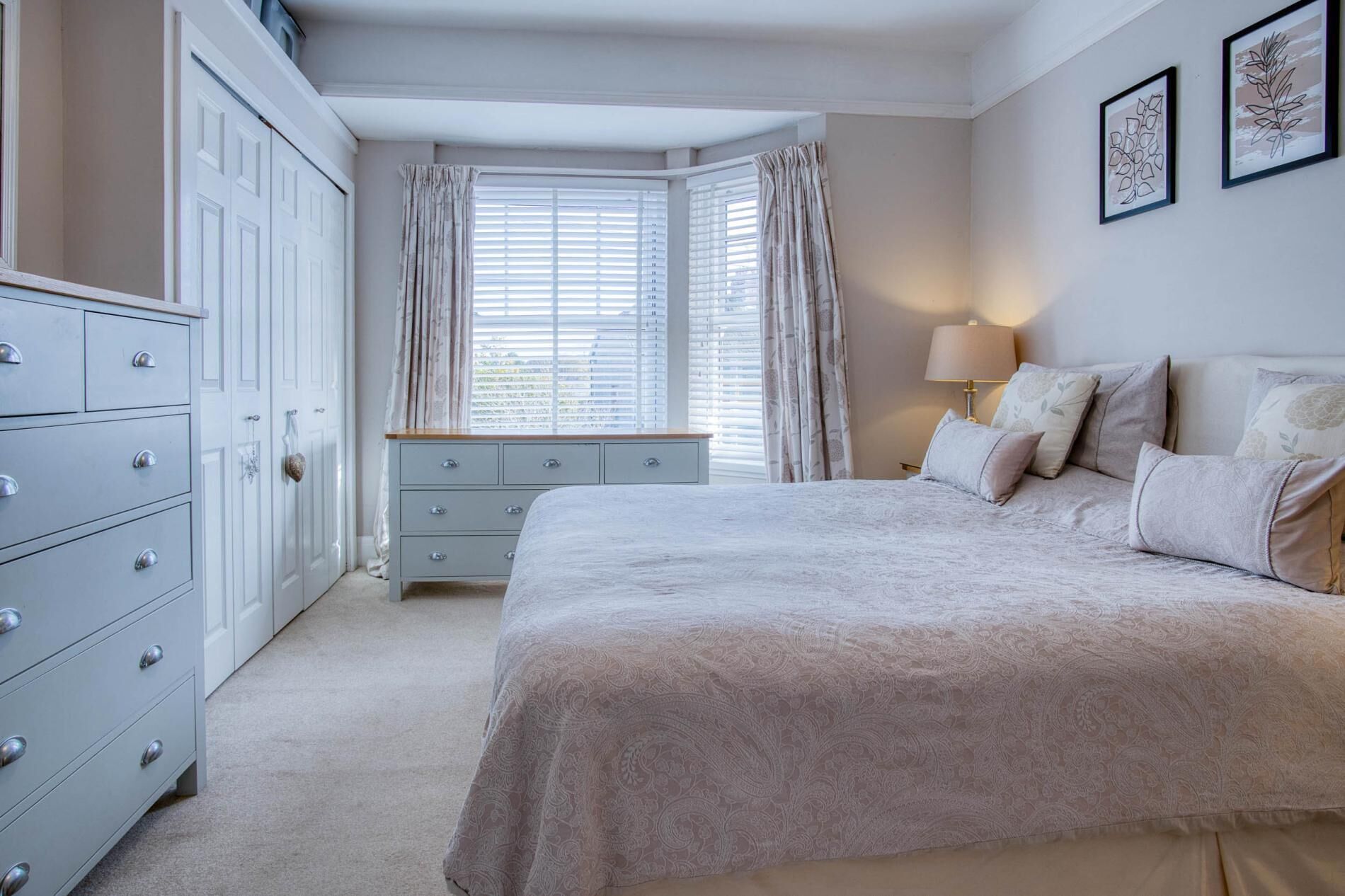 Een slaapkamer met een groot bed, een kaptafel, een raam met gordijnen en een wandlamp.