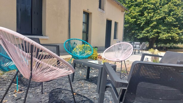 An outdoor seating area with chairs and a table, a building with windows, and a tree in the background. - Country cottage, Verette cottage (Écueillé)